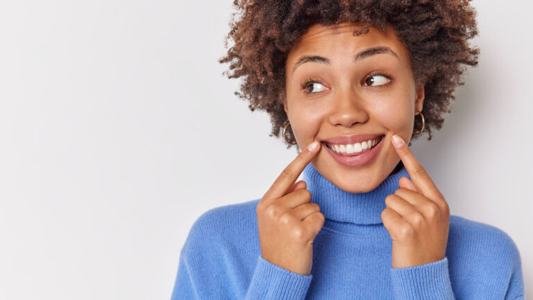 Positive carefree woman smiles broadly points at her perfect even teeth indicates at corners of lips looks away wears casual blue jumper isolated over white background with blank copy space